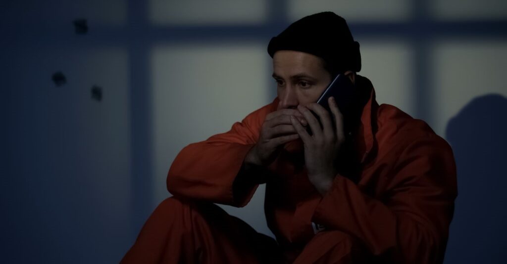 A prisoner behind bars in an orange jump suit whispering on a mobile phone