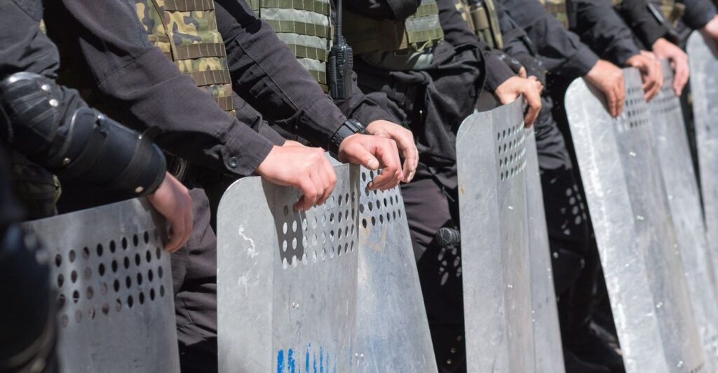 A line of police officers in riot gear holding large metal shields